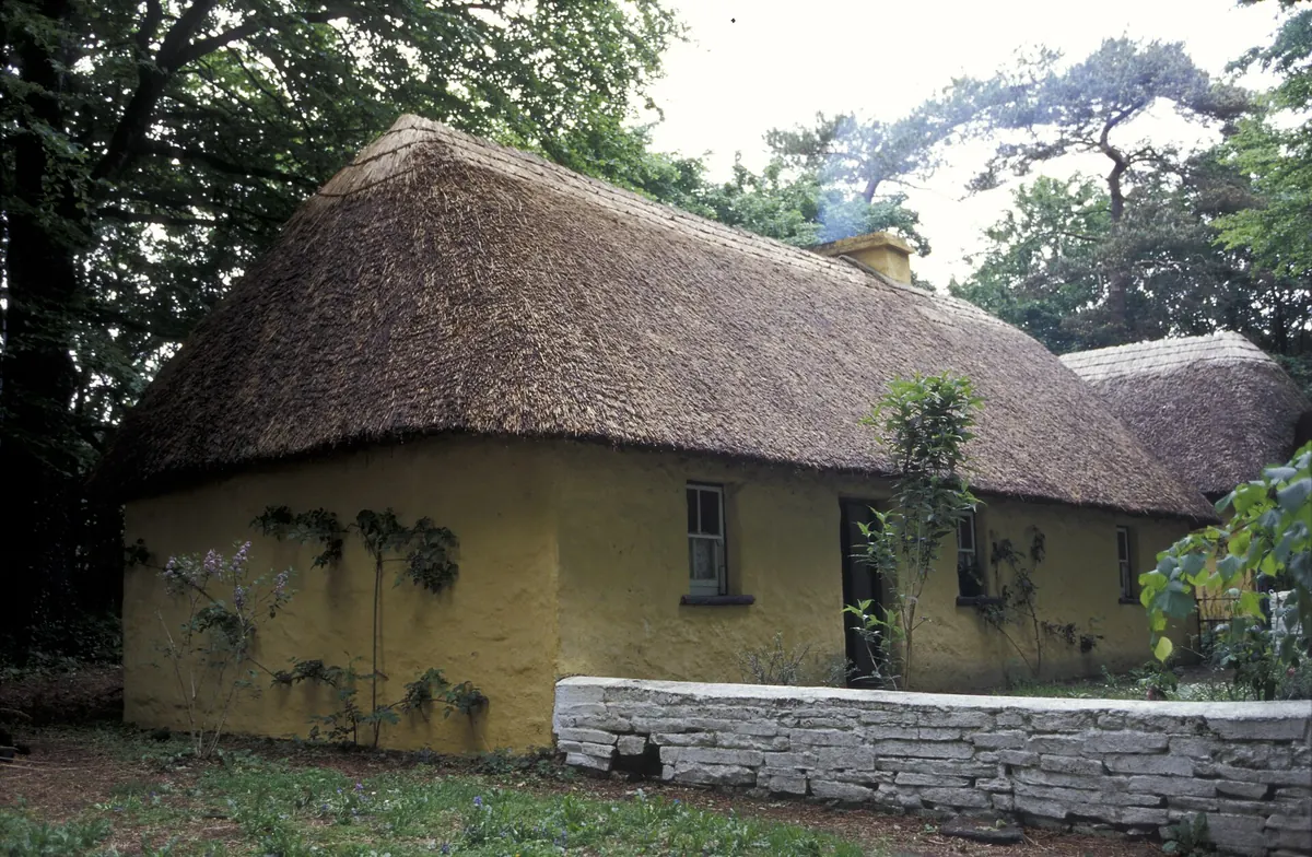 Un cottage dal tetto di paglia dalle pareti gialle nel Bunratty Folk Park, con un basso muro in pietra davanti e alberi alle spalle