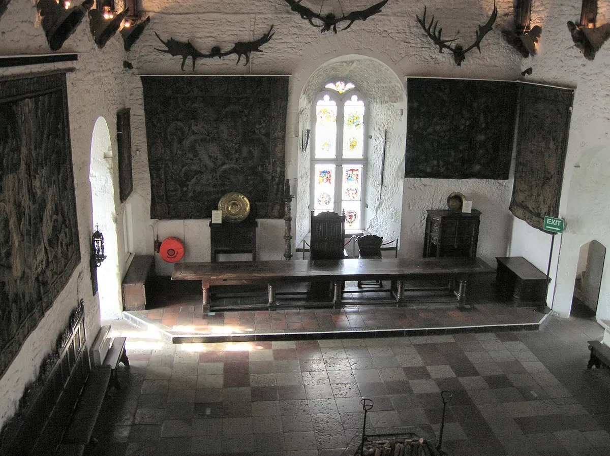 La Great Hall du château de Bunratty en plein jour, une longue table en bois sur une estrade sous des tapisseries et un vitrail, avec des bois de cerf sur les murs blanchis à la chaux