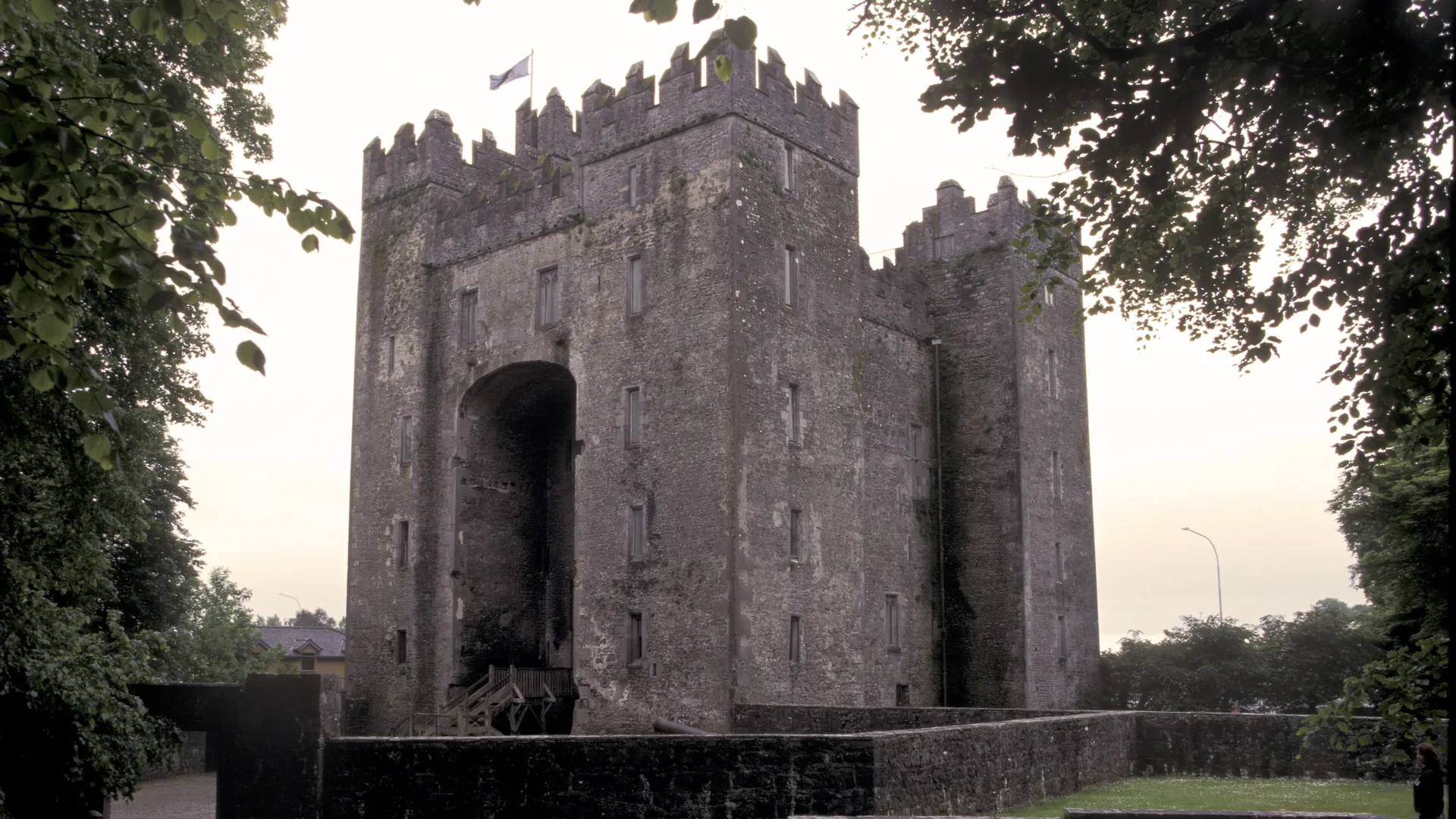 Bunratty Castle in der Abenddämmerung, ein grauer steinerner Wohnturm mit Zinnen und einer hohen bogenförmigen Nische, umrahmt von dunklen Bäumen, mit einer wehenden Flagge über dem Dach
